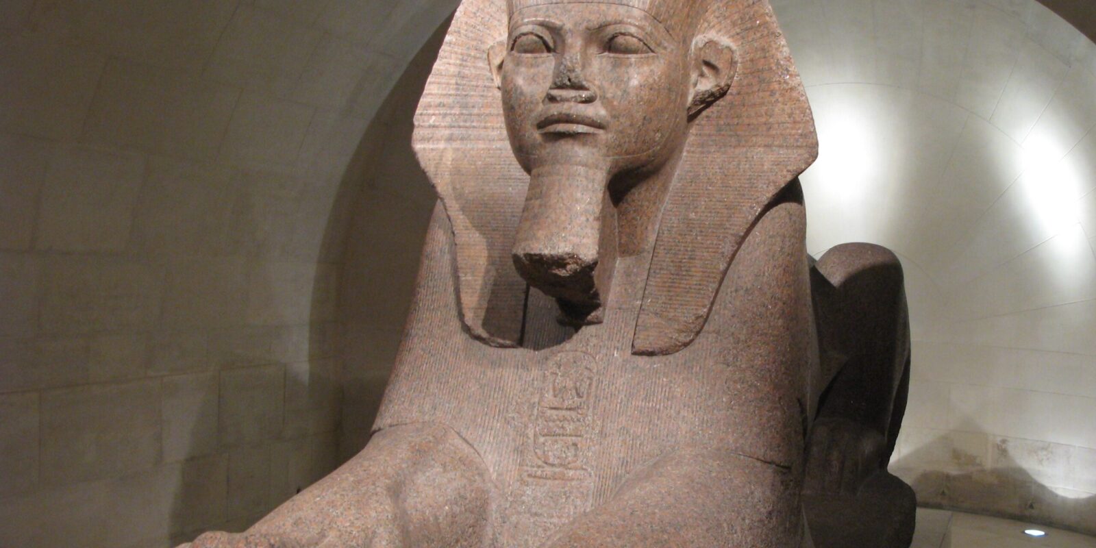 Large red granite statue of a Pharaoh wearing a nemes headdress with a beard, seated in a museum gallery.