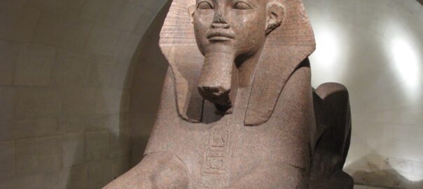 Large red granite statue of a Pharaoh wearing a nemes headdress with a beard, seated in a museum gallery.