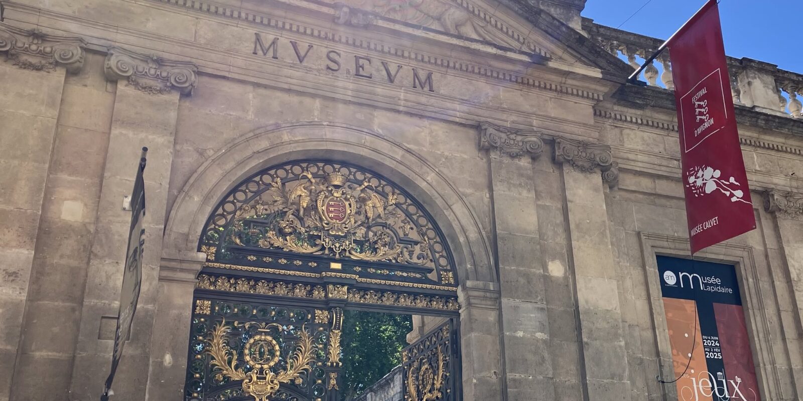 Stone building entrance with ornate wrought-iron gate featuring gold embellishments and a crest, flanked by red festival banners; sun glare at top of facade.