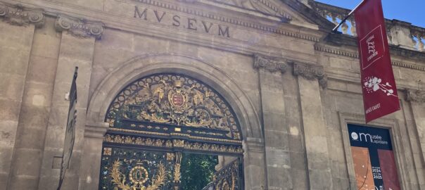 Stone building entrance with ornate wrought-iron gate featuring gold embellishments and a crest, flanked by red festival banners; sun glare at top of facade.